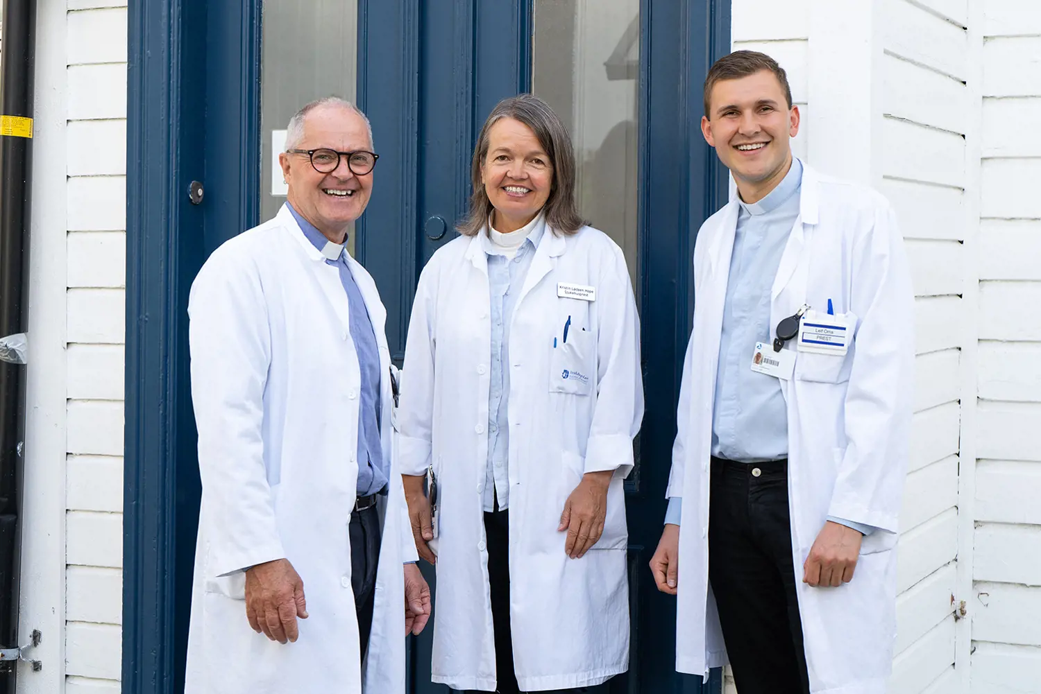 Tre smilende sykehusprester i hvite frakker står foran en blå dør